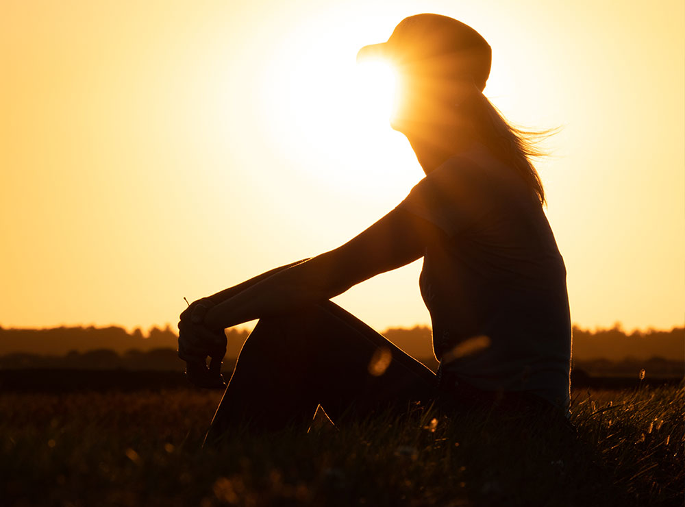 Una silueta de una mujer con una gorra de béisbol, sentada en un campo al atardecer.